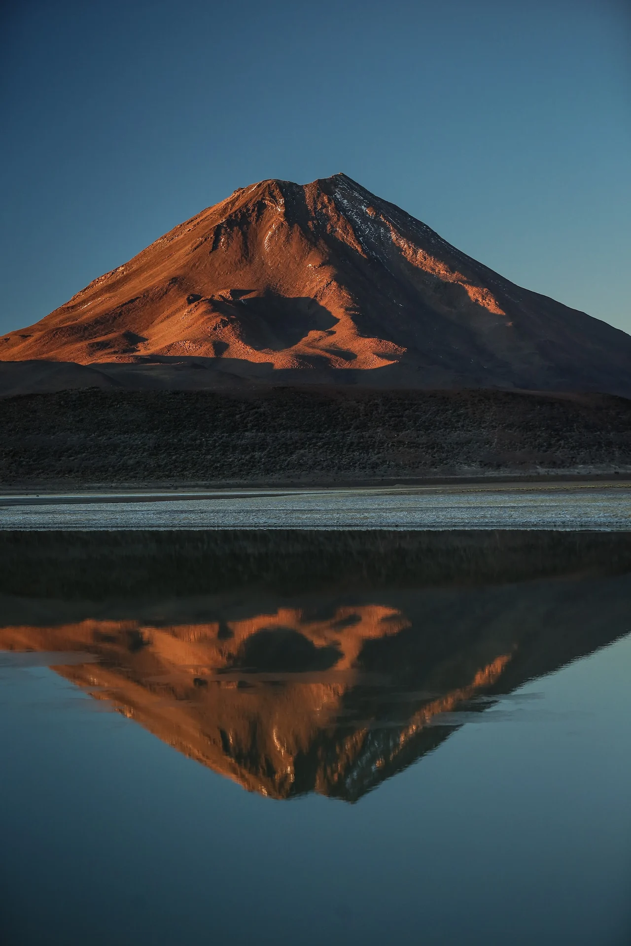 Sunlit mountain in Atacama Desert reflected in calm lake at golden hour under clear blue sky.