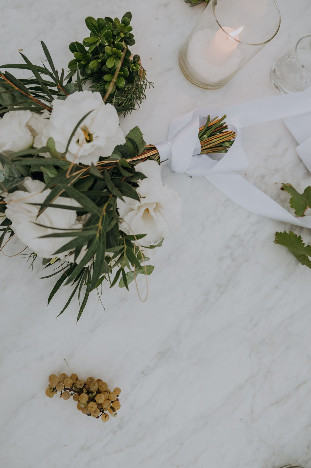 Elegant marble table setting with white flower bouquet, green grapes, lit candle in glass holder, and potted plant.