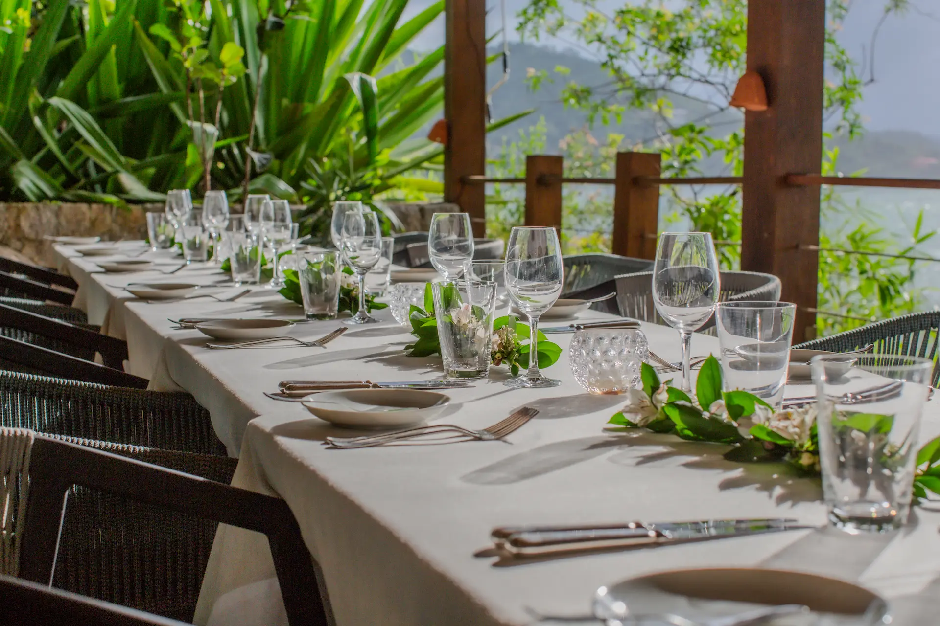 Elegant outdoor dining table with white linens and floral centerpieces, set in a tropical garden overlooking water.