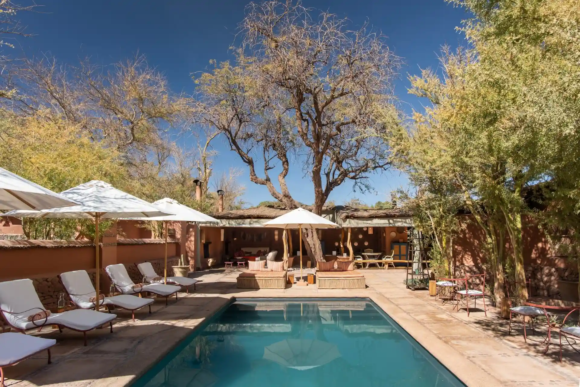 Outdoor pool area with cushioned lounge chairs, white umbrellas, rustic patio, and clear blue sky.